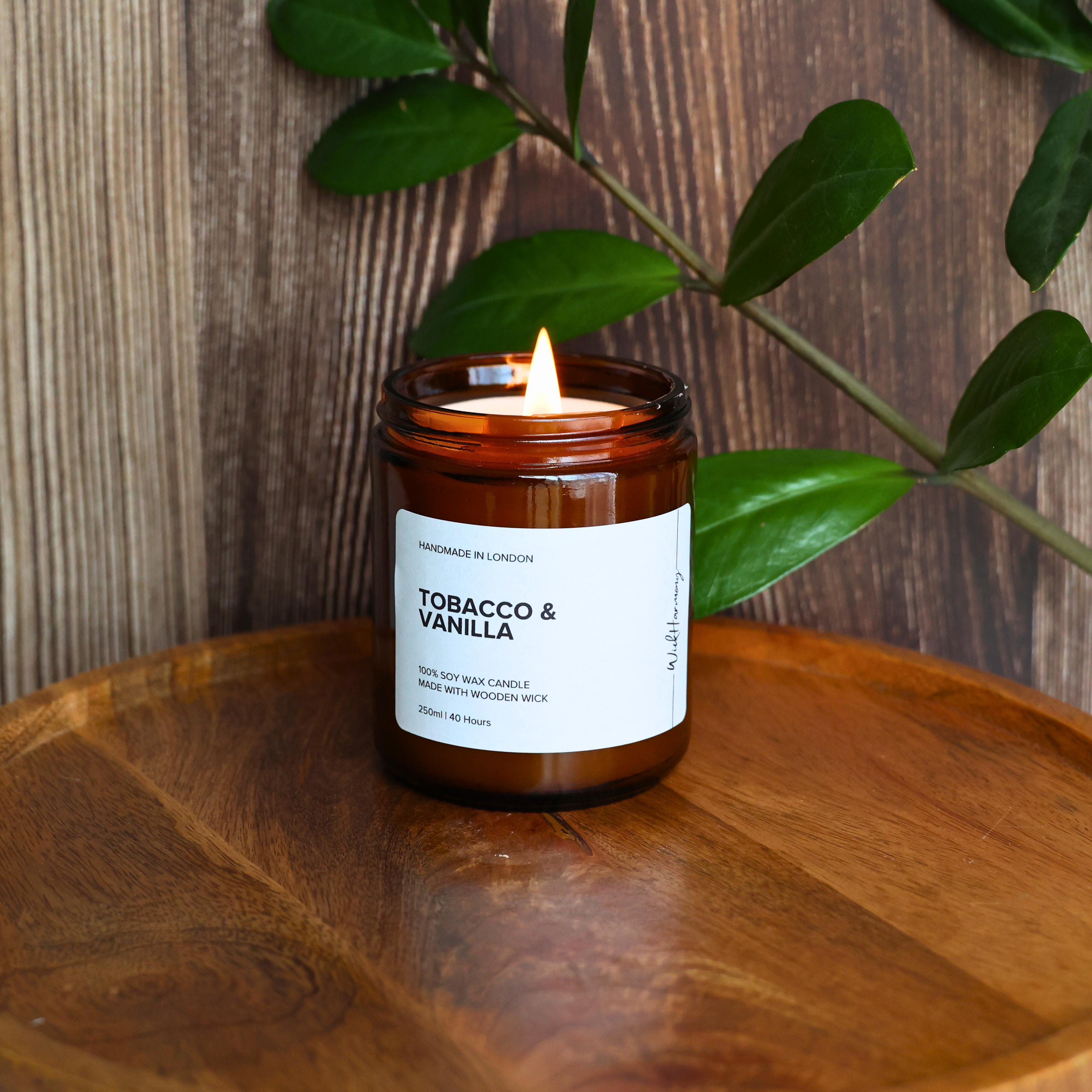 The 250ml 'Tobacco & Vanilla' wooden wick candle burning with a gentle flame, set on a wooden tray with a dark wood background.