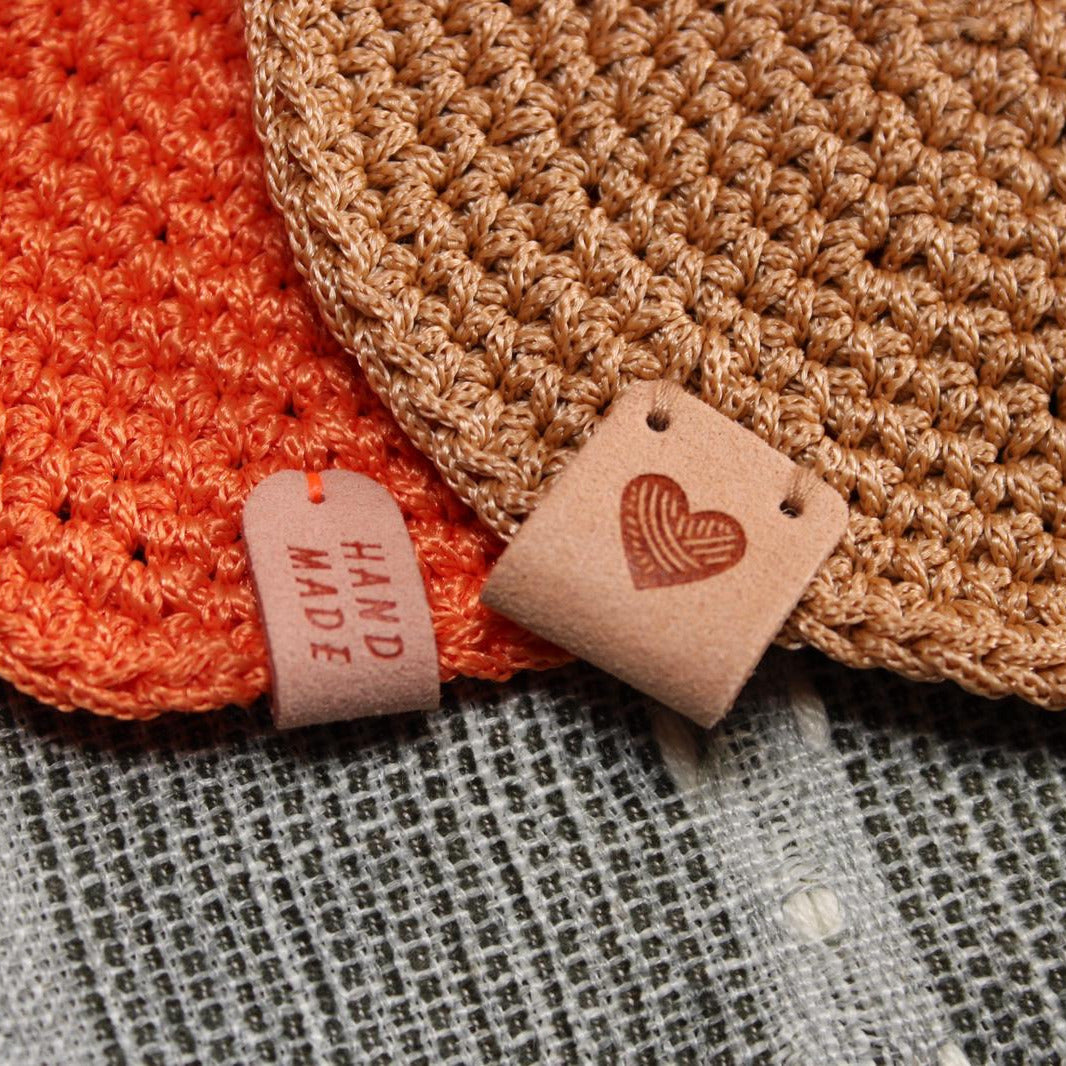 A close-up of the charming leatherette tags on the WickHarmony crochet candle coasters, one reading "Hand Made" and the other with a heart