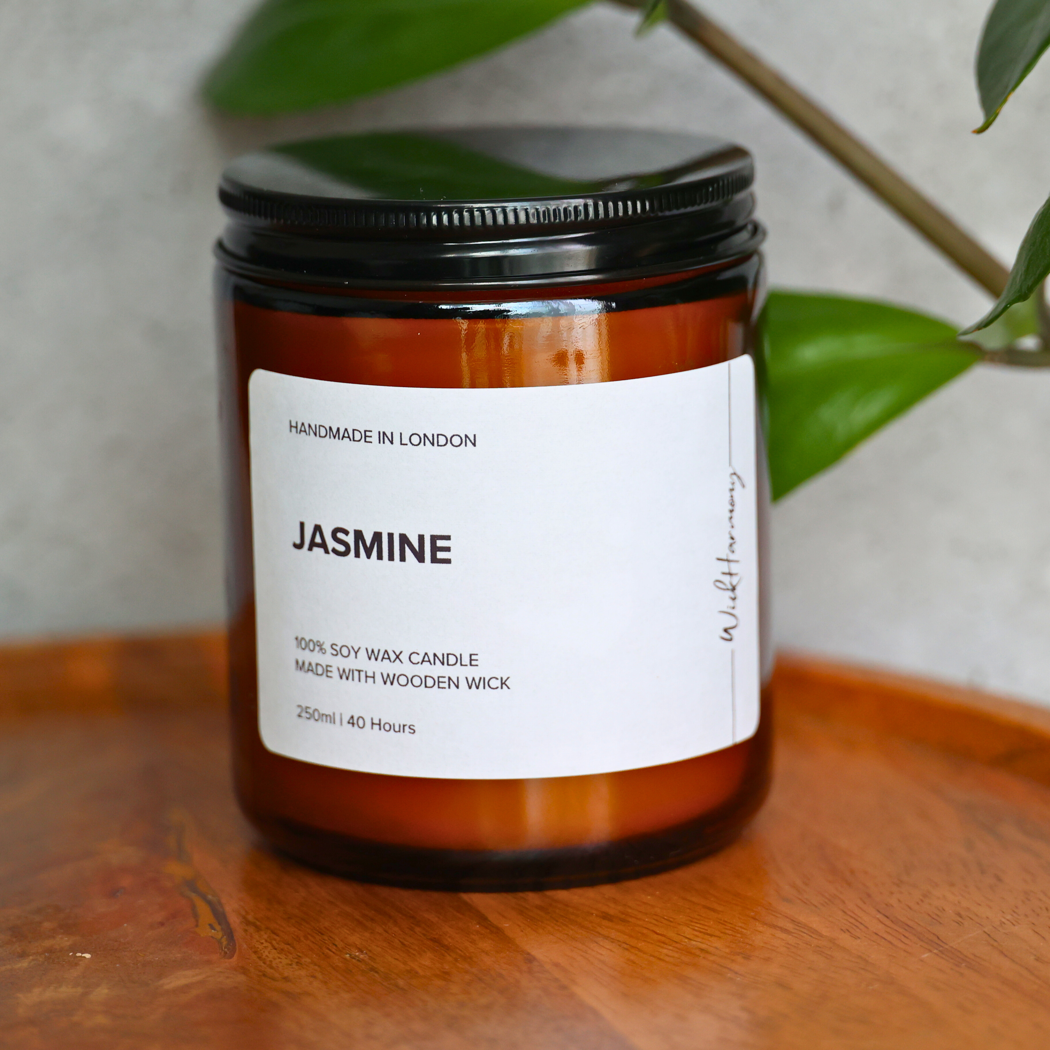 A 250ml amber jar 'Jasmine' soy wax candle with a black lid, sitting on a wooden tray in front of a green plant.