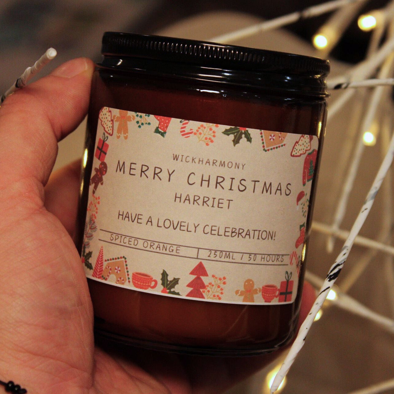 Hand holding a large 250ml personalised 'Merry Christmas Harriet' candle with festive fairy lights in the background.