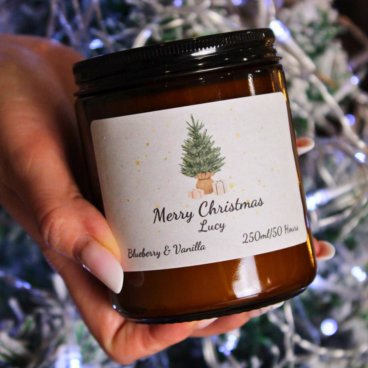 A hand holding a large 250ml candle personalised with the name 'Lucy' against a background of blue and white Christmas lights