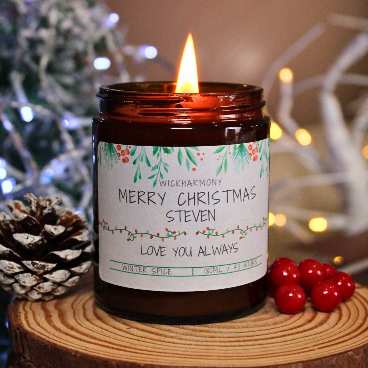 A lit 180ml Winter Spice candle personalised with 'Merry Christmas Steven' next to a pinecone, creating a warm, festive glow.