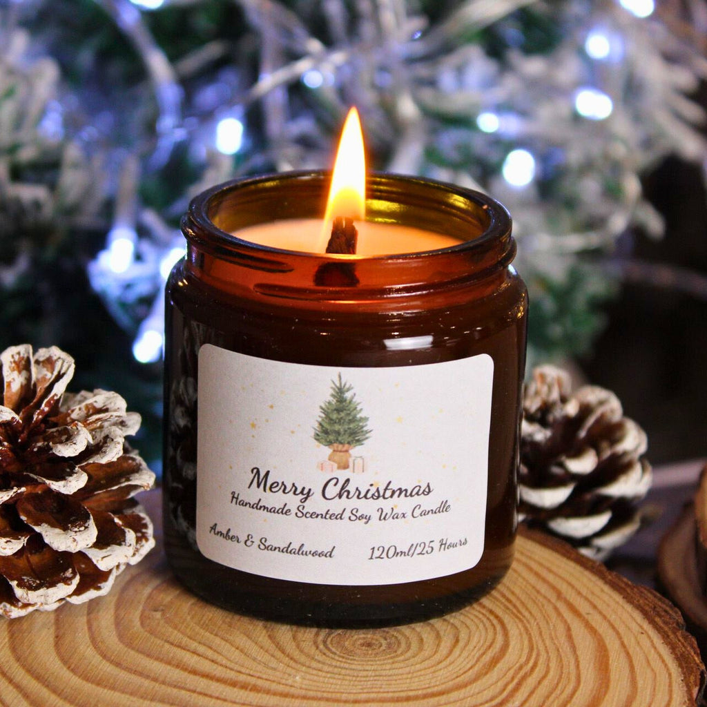 A lit personalised soy wax candle with a Christmas tree label, sitting on a wood slice between two snowy pinecones.