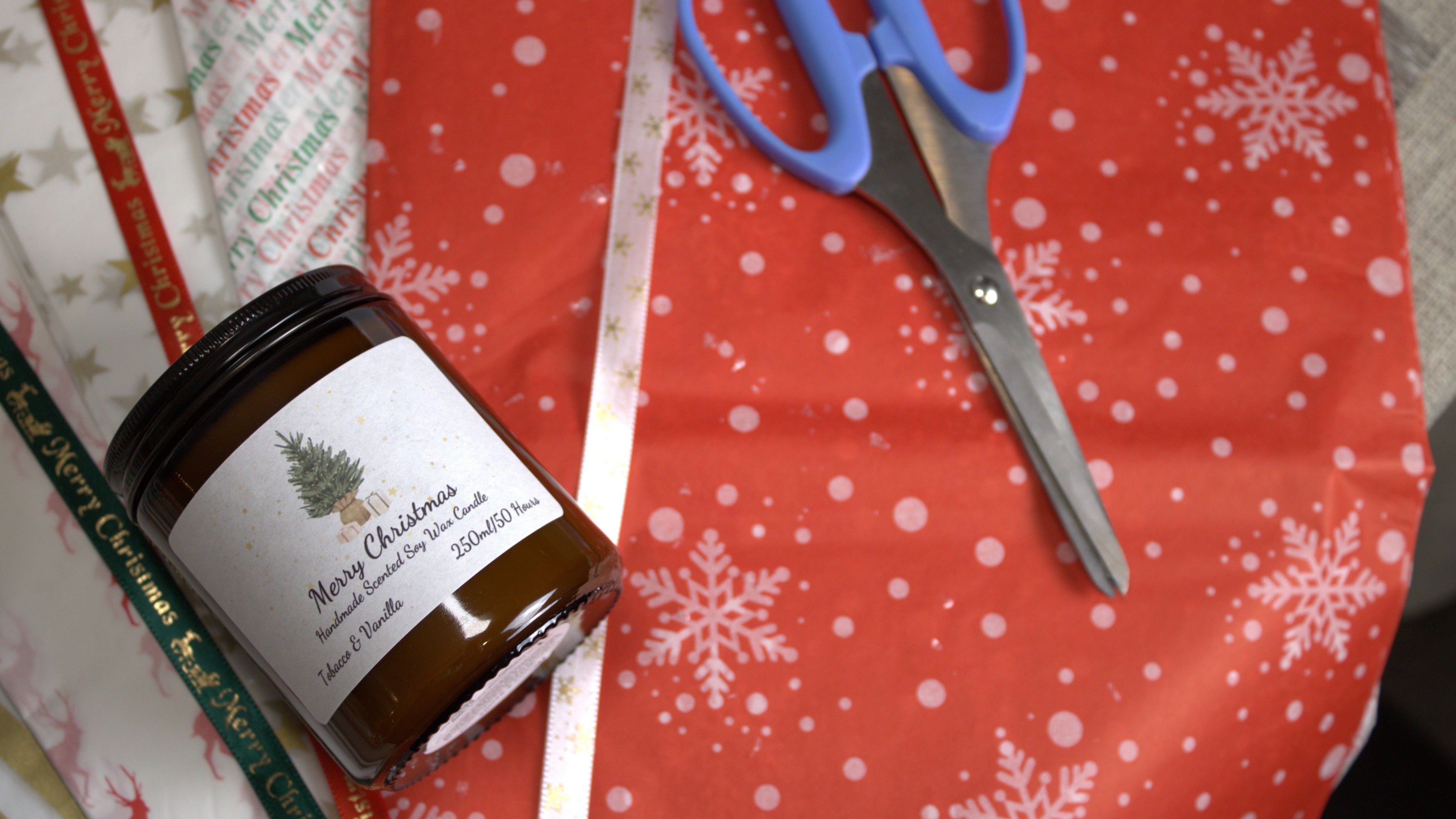A 250ml 'Merry Christmas' amber jar candle with 'Tobacco & Vanilla' scent, shown in a festive gift-wrapping scene with scissors and holiday paper.