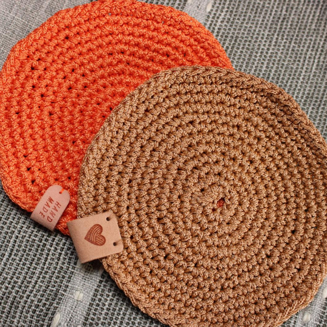 A pair of handmade crochet candle coasters by WickHarmony in rustic orange and tan brown, shown on a linen surface
