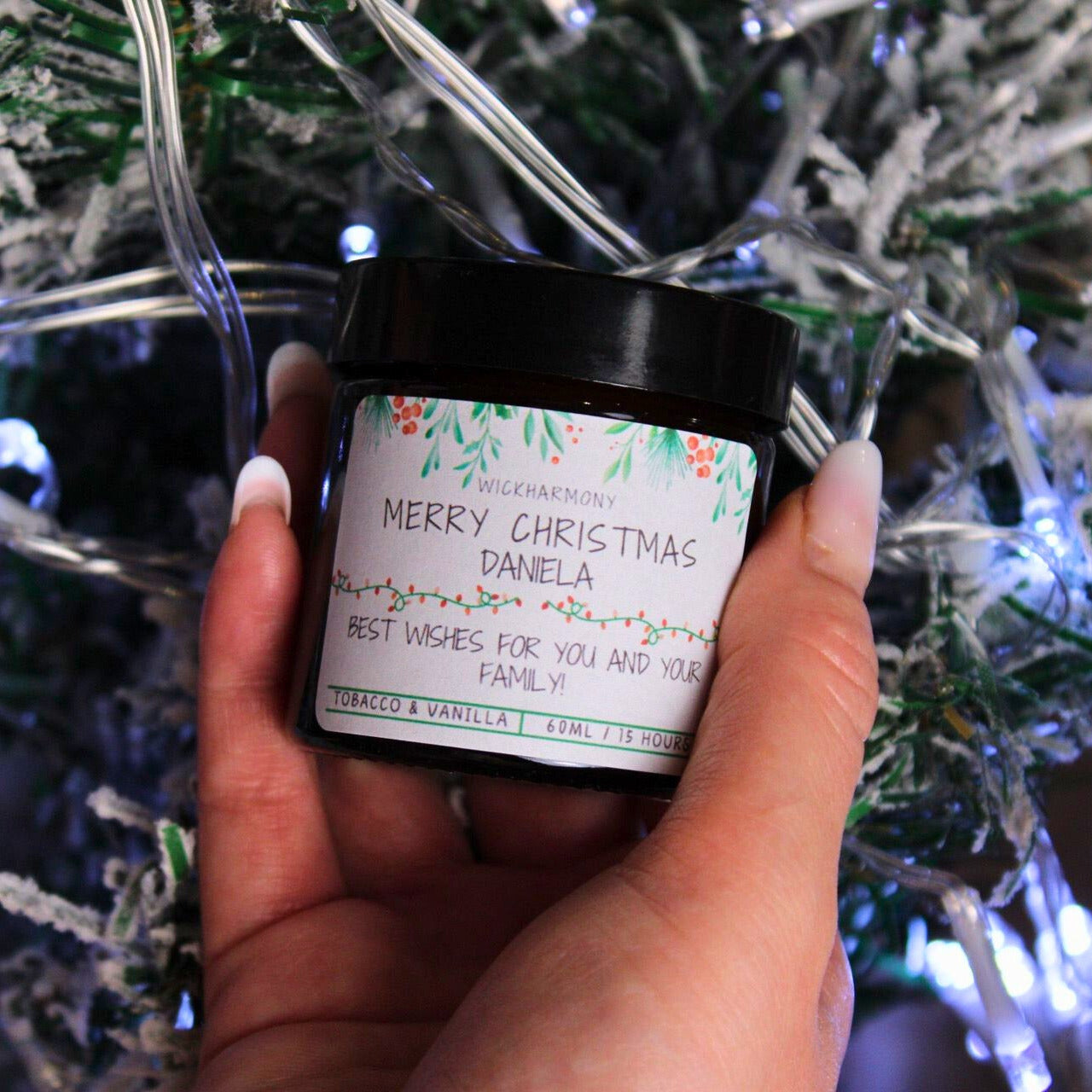 Hand holding a small personalised 'Merry Christmas Daniela' soy candle in front of a festive Christmas tree