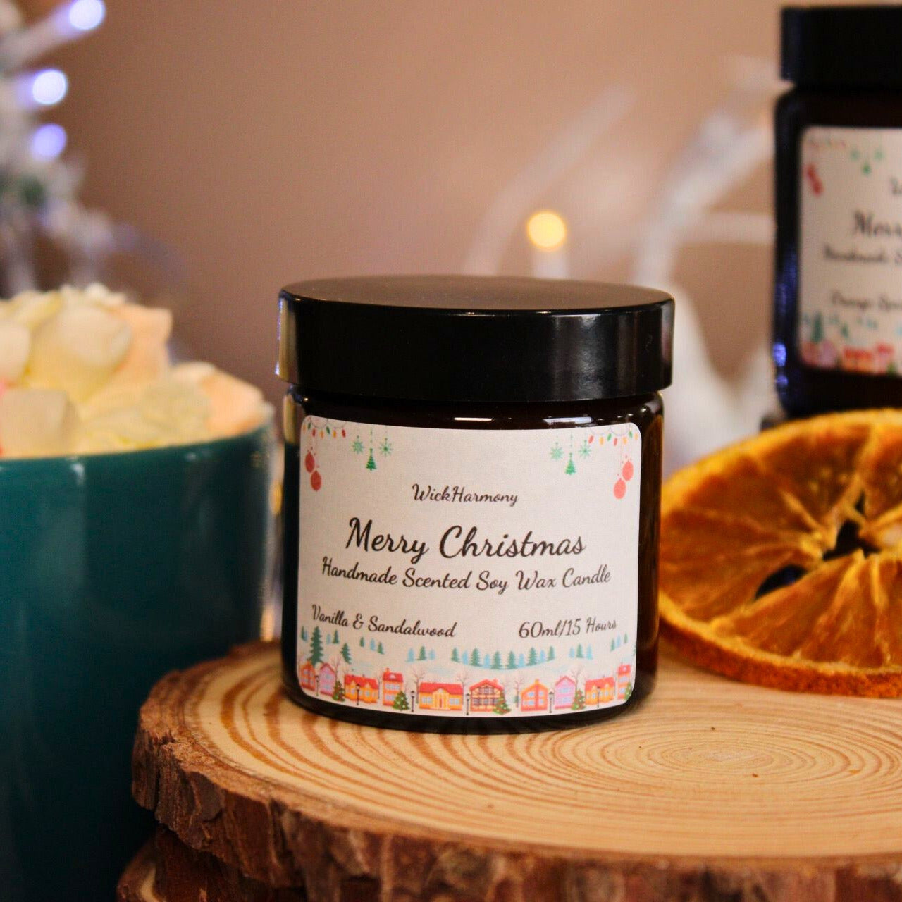A small 60ml Christmas village soy candle on a wood slice next to a dried orange and a mug of hot chocolate.