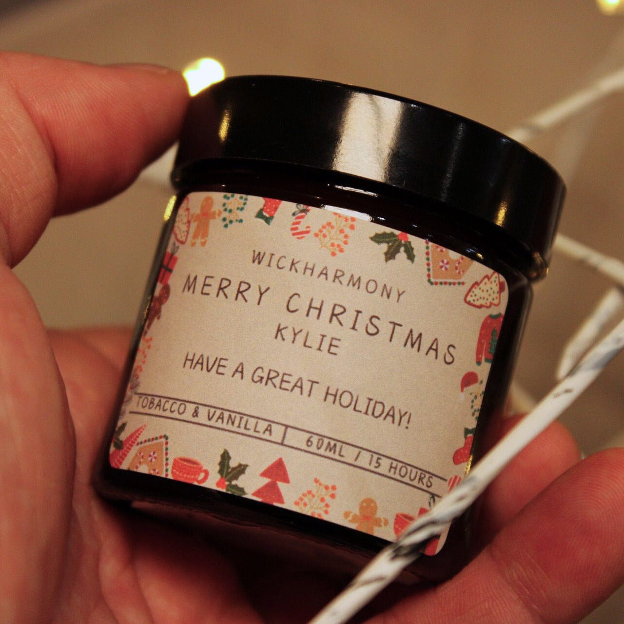 A close-up of a hand holding a small 60ml personalised 'Merry Christmas Kylie' candle with a rustic label design.