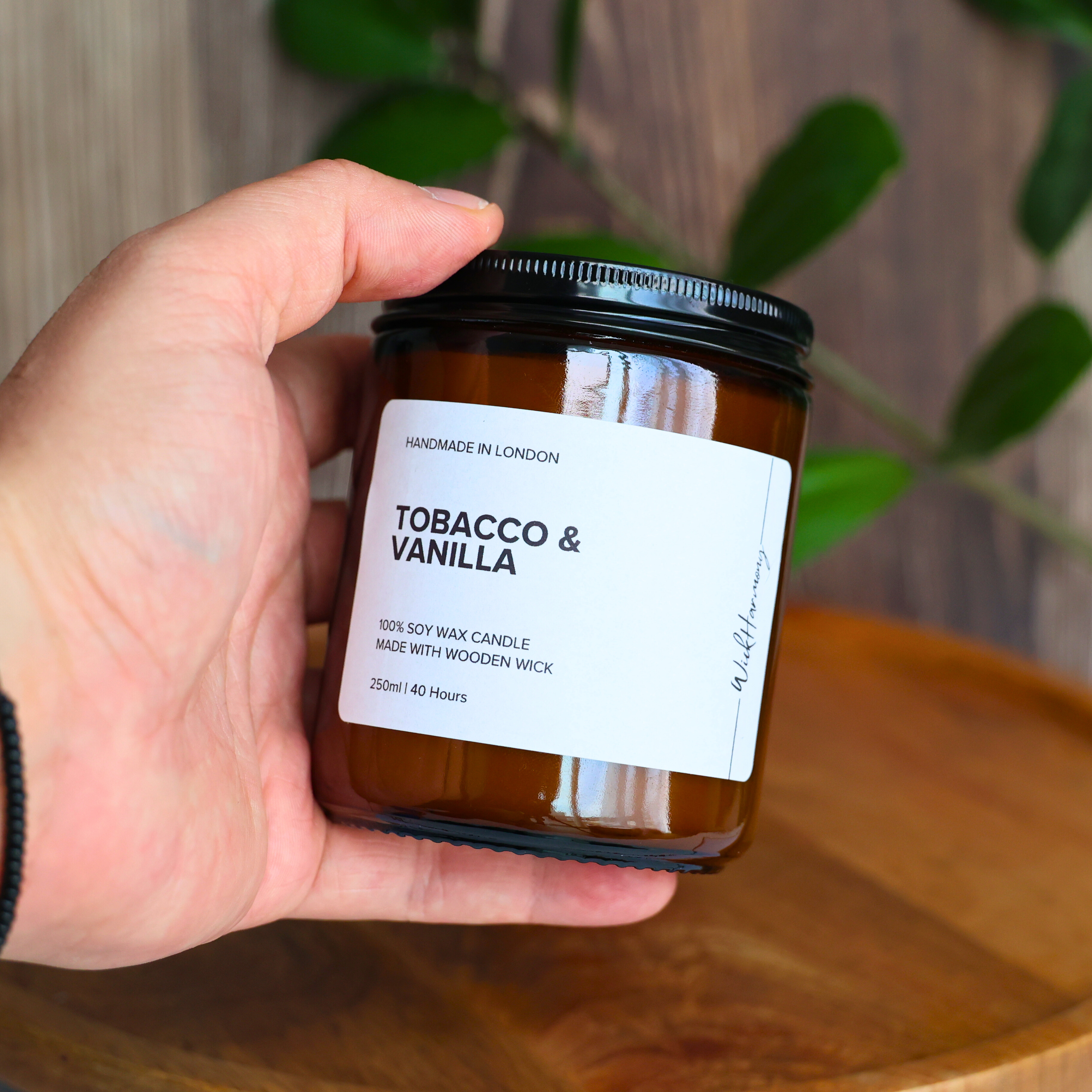A hand holding the 250ml amber jar 'Tobacco & Vanilla' soy wax candle by WickHarmony to demonstrate its size.