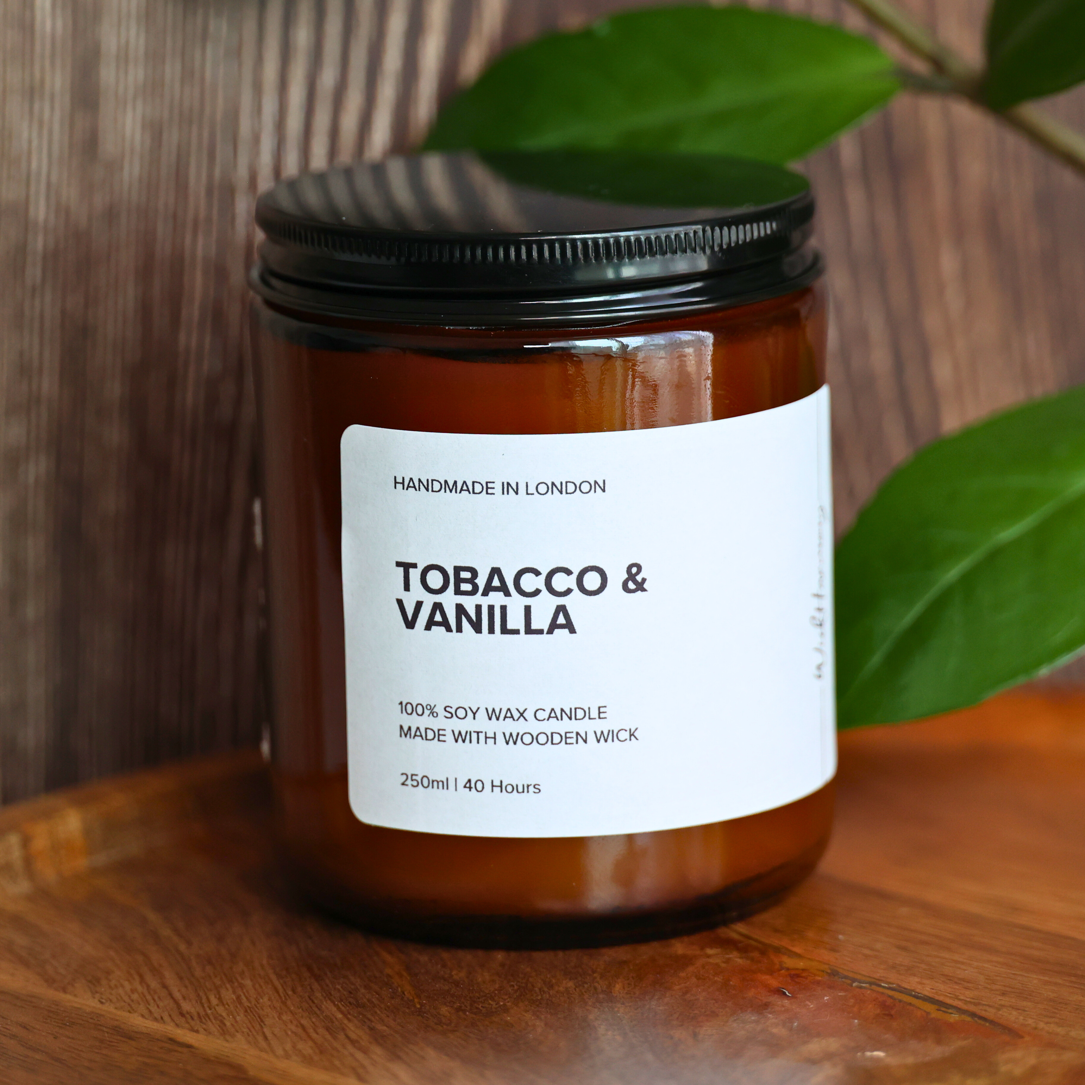 A 250ml 'Tobacco & Vanilla' soy wax candle in an amber jar, shown on a wooden tray with a rustic wood and green plant background.