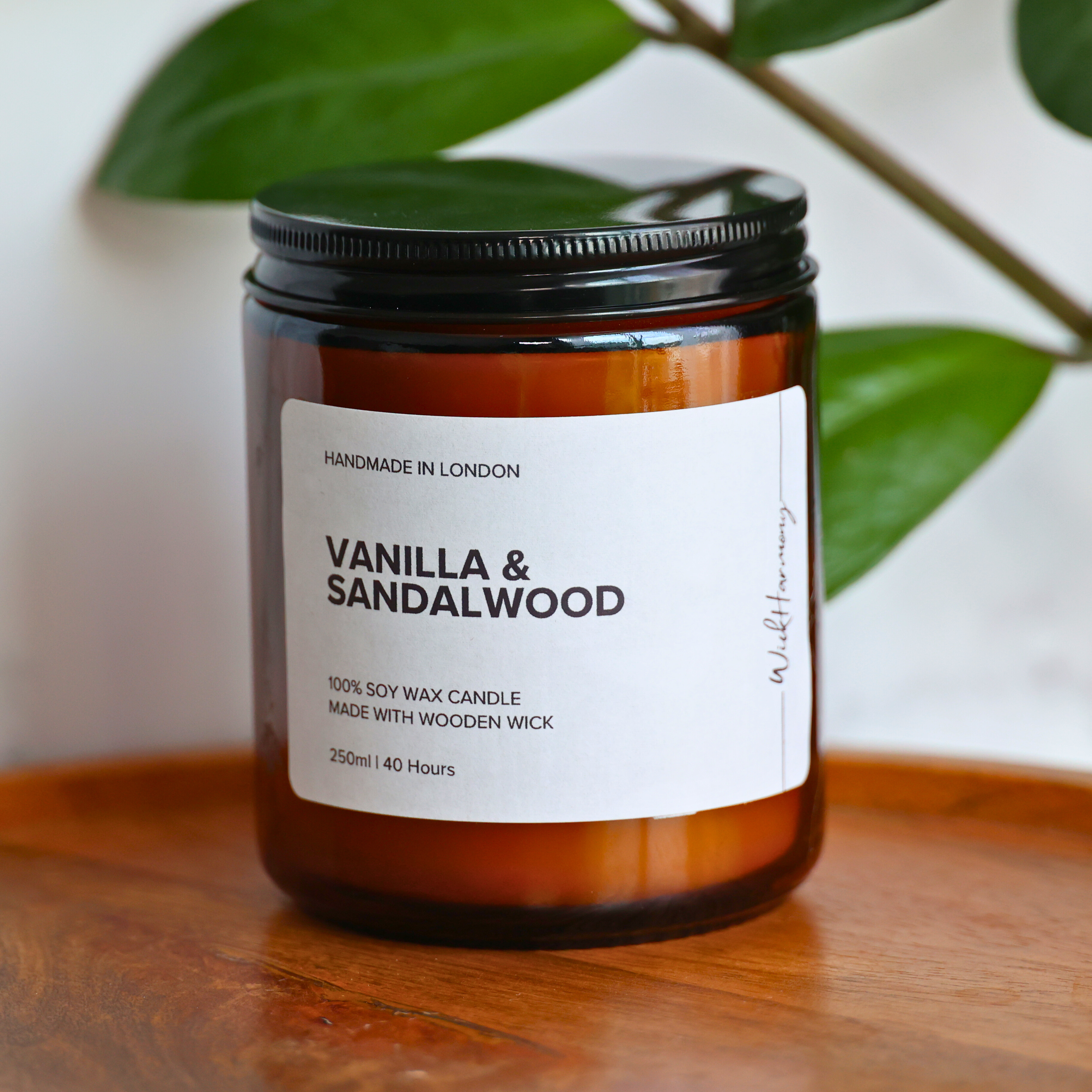 A 250ml amber jar 'Vanilla & Sandalwood' soy candle with a black lid, sitting on a wooden tray with a green plant in the background.