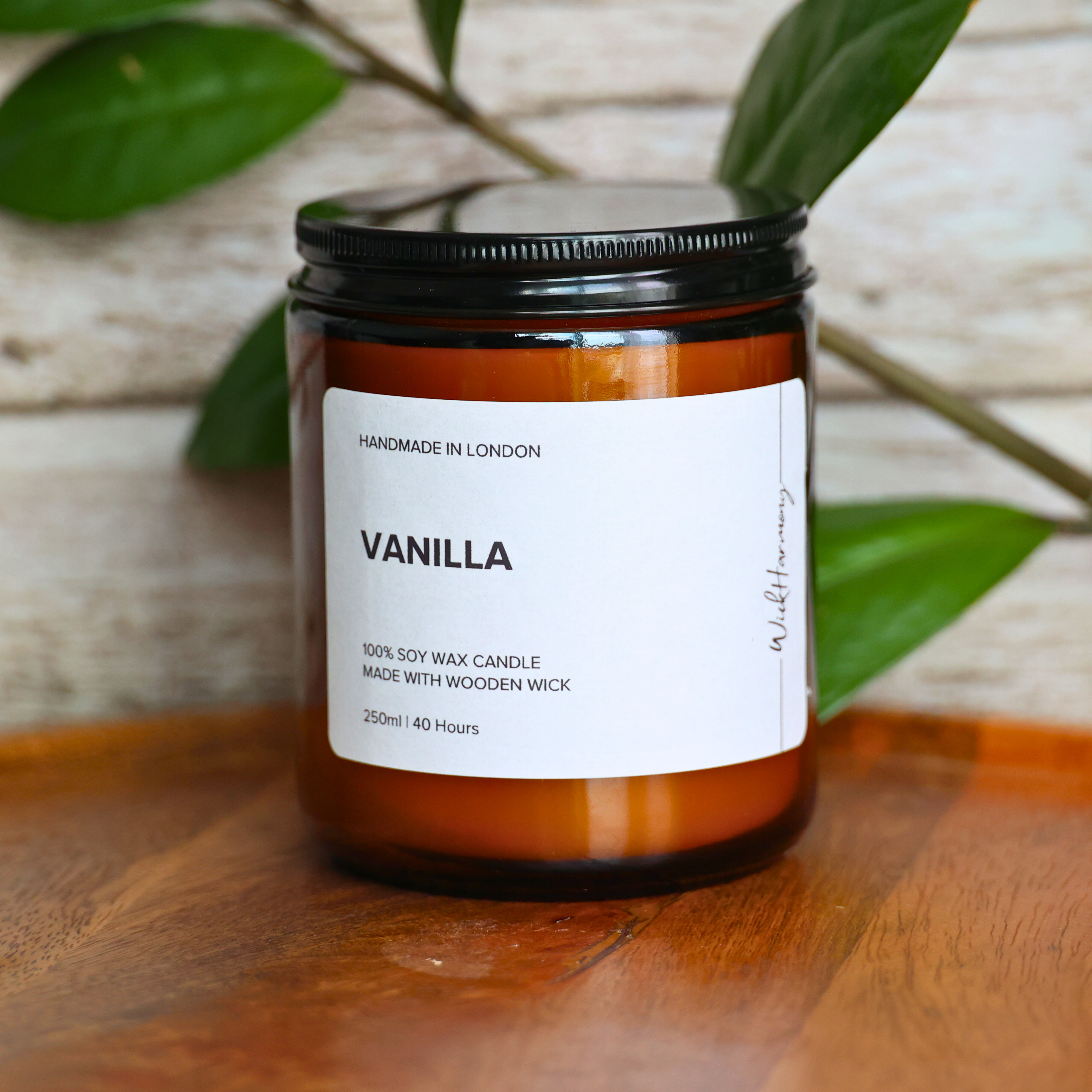 A 250ml 'Vanilla' soy wax candle by WickHarmony, shown in an amber jar on a wooden tray with a rustic wood and plant background